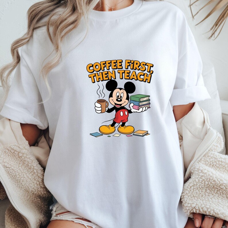 May include: White t-shirt with a cartoon Mickey Mouse design. Mickey is holding a coffee cup and books. The text "COFFEE FIRST THEN TEACH" is printed above the character in orange and yellow.