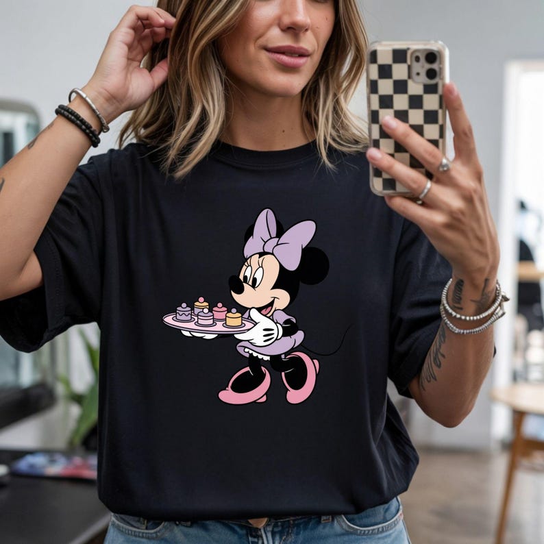 Puede incluir: Camiseta negra con un dise&ntilde;o de Minnie Mouse. Minnie aparece con un vestido y lazo morados, sosteniendo una bandeja con peque&ntilde;os pasteles. El dise&ntilde;o incluye detalles en rosa en sus zapatos y la bandeja. La camiseta es de estilo casual.