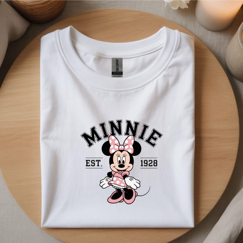 Może przedstawiać: Biała koszulka z grafiką Myszki Minnie w r&oacute;żowej sukience w kropki i kokardą. Słowo "MINNIE" jest wygięte nad postacią, z napisem "EST. 1928" poniżej. Koszulka jest złożona na drewnianej powierzchni.