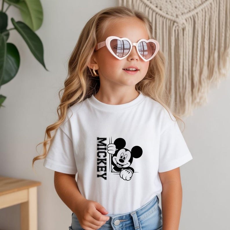 May include: White t-shirt featuring a black and white graphic of Mickey Mouse peeking out from behind a rectangle with the word "Mickey" printed vertically. The shirt is worn by a young person wearing heart-shaped sunglasses.