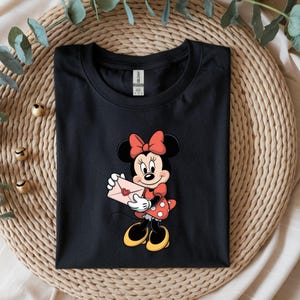 Op de afbeelding: Zwart T-shirt met een cartoon van Minnie Mouse die een roze envelop met een hart vasthoudt. Minnie Mouse draagt een rode strik, een rode jurk met witte stippen en gele schoenen. Het shirt is opgevouwen op een geweven mat.