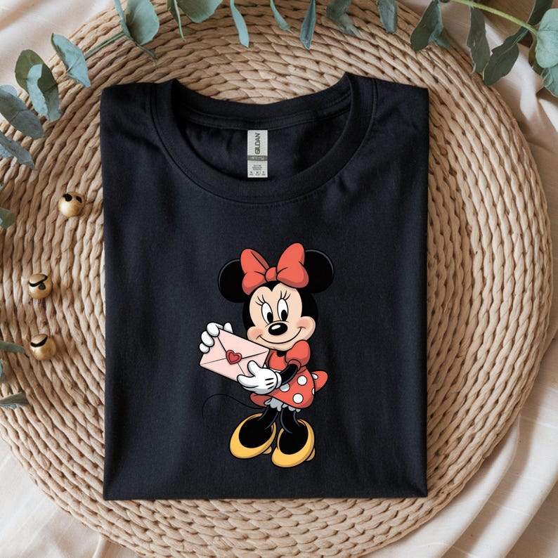 Op de afbeelding: Zwart T-shirt met een cartoon van Minnie Mouse die een roze envelop met een hart vasthoudt. Minnie Mouse draagt een rode strik, een rode jurk met witte stippen en gele schoenen. Het shirt is opgevouwen op een geweven mat.