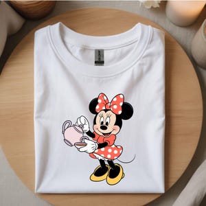 Puede incluir: Camiseta blanca con un dise&ntilde;o de Minnie Mouse sosteniendo una tetera y una taza rosas. Minnie lleva un vestido rojo con lunares blancos, un lazo a juego y zapatos amarillos. La camiseta est&aacute; doblada sobre una superficie de madera.