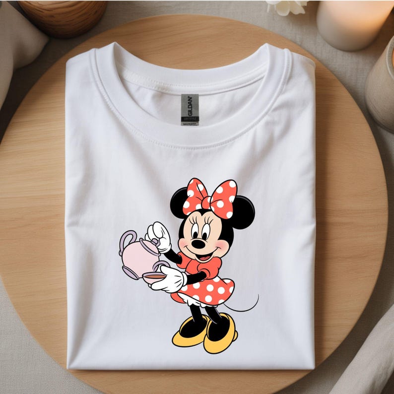 Puede incluir: Camiseta blanca con un dise&ntilde;o de Minnie Mouse sosteniendo una tetera y una taza rosas. Minnie lleva un vestido rojo con lunares blancos, un lazo a juego y zapatos amarillos. La camiseta est&aacute; doblada sobre una superficie de madera.