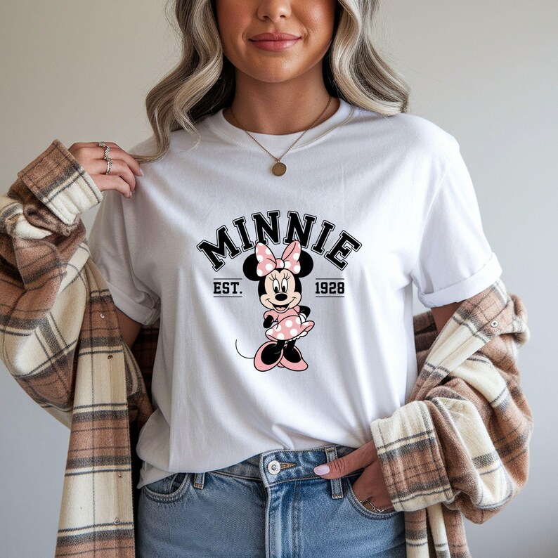 Może przedstawiać: Biała koszulka z grafiką Myszki Minnie. Projekt zawiera napis "Minnie" w czcionce uniwersyteckiej, tekst "EST. 1928" oraz kresk&oacute;wkowy wizerunek Myszki Minnie w r&oacute;żowej sukience i kokardce.