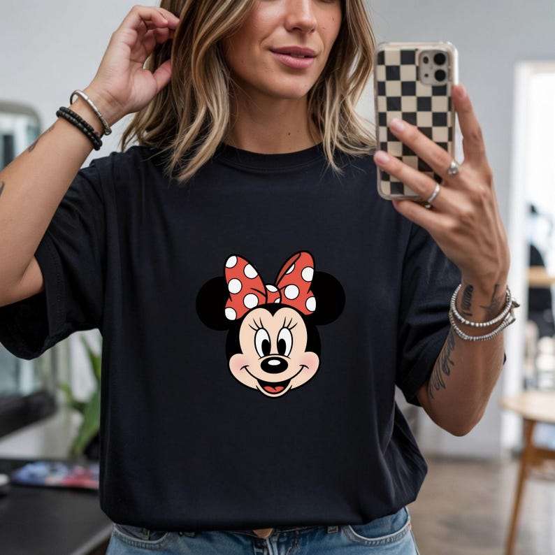 May include: Black t-shirt featuring a Minnie Mouse graphic. The graphic shows Minnie's face with a red bow with white polka dots. The shirt is a solid black color.