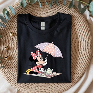 Puede incluir: Camiseta negra con un dise&ntilde;o de dibujos animados de Minnie Mouse. Minnie aparece sosteniendo un paraguas de lunares, participando en una fiesta de t&eacute; con una tetera y tazas. El dise&ntilde;o incluye detalles en rosa, blanco y amarillo.