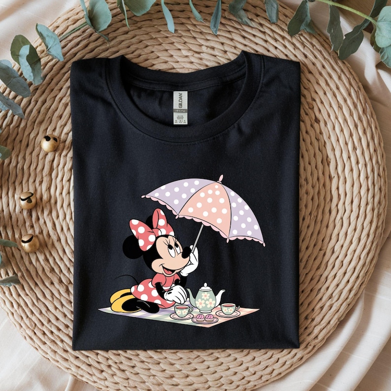 Puede incluir: Camiseta negra con un dise&ntilde;o de dibujos animados de Minnie Mouse. Minnie aparece sosteniendo un paraguas de lunares, participando en una fiesta de t&eacute; con una tetera y tazas. El dise&ntilde;o incluye detalles en rosa, blanco y amarillo.
