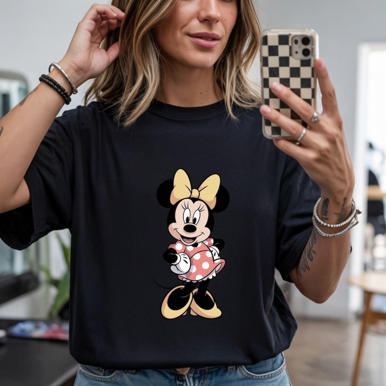 Op de afbeelding: Zwart T-shirt met een Minnie Mouse-ontwerp. Het stripfiguur draagt een roze jurk met stippen en een gele strik. Het T-shirt heeft een casual stijl.