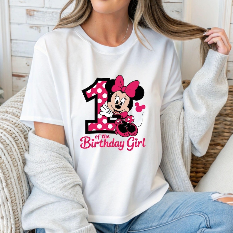 May include: White t-shirt featuring a graphic of Minnie Mouse with a pink polka-dotted number 1 and the text "of the Birthday Girl" in pink. The design includes a pink bow and shoes.