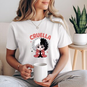 以下が含まれることがあります： クルエラ・ド・ヴィルの漫画イラストが描かれた白いTシャツ。キャラクターは白と黒の髪で、赤いコートを着ています。「クルエラ」の文字がピンク色でキャラクターの上にプリントされています。Tシャツはカジュアルなスタイルです。