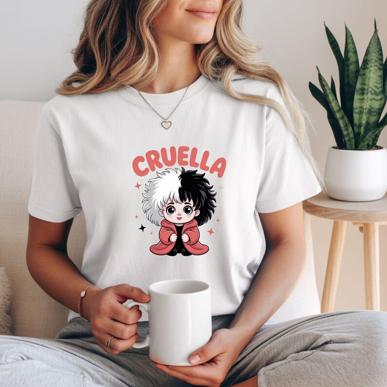 以下が含まれることがあります： クルエラ・ド・ヴィルの漫画イラストが描かれた白いTシャツ。キャラクターは白と黒の髪で、赤いコートを着ています。「クルエラ」の文字がピンク色でキャラクターの上にプリントされています。Tシャツはカジュアルなスタイルです。