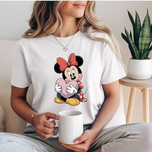 Op de afbeelding: Wit T-shirt met een cartoon Minnie Mouse die een roze hart vasthoudt. Minnie Mouse heeft een rode strik, een rood-wit gestippelde jurk en gele schoenen. Het T-shirt heeft een casual stijl.