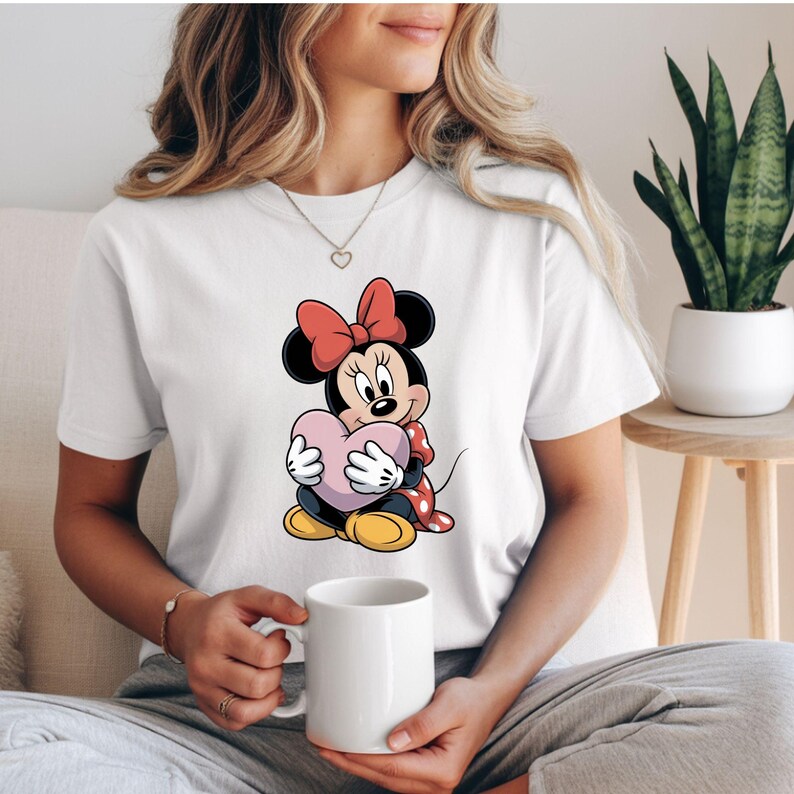 Op de afbeelding: Wit T-shirt met een cartoon Minnie Mouse die een roze hart vasthoudt. Minnie Mouse heeft een rode strik, een rood-wit gestippelde jurk en gele schoenen. Het T-shirt heeft een casual stijl.