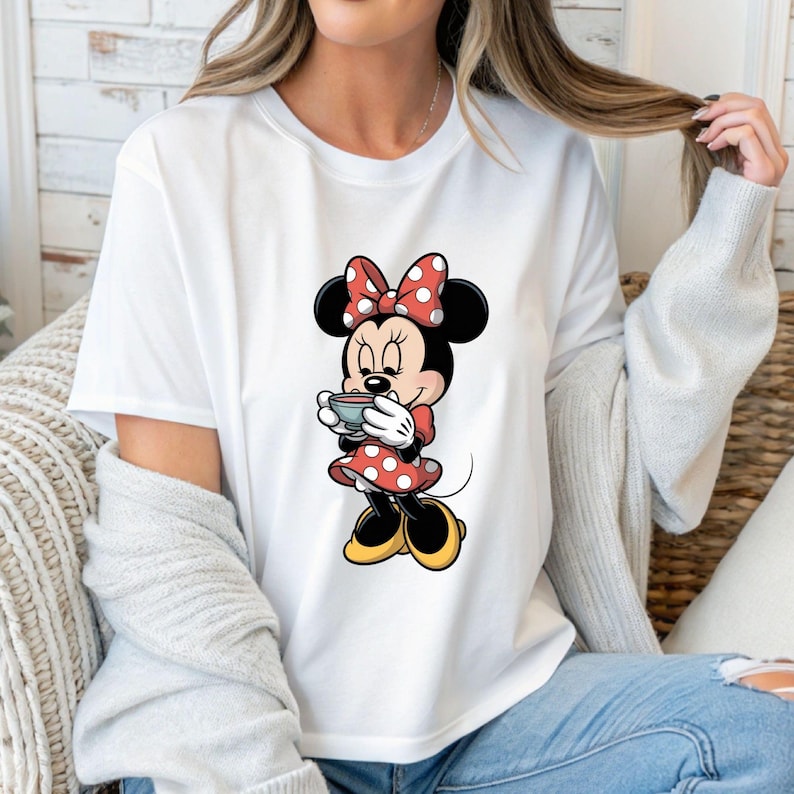 Puede incluir: Camiseta blanca con una ilustraci&oacute;n de dibujos animados de Minnie Mouse sosteniendo una taza. Minnie Mouse lleva un vestido rojo con lunares blancos, un lazo a juego y zapatos amarillos. El dise&ntilde;o est&aacute; centrado en la camiseta.