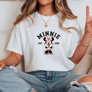 Może przedstawiać: Biała koszulka z grafiką Myszki Minnie. Projekt zawiera napis "MINNIE" w czarnym, łukowatym tekście nad kresk&oacute;wkowym wizerunkiem Myszki Minnie, z napisem "EST. 1928" poniżej. Myszka Minnie ma na sobie r&oacute;żową sukienkę w kropki i kokardę.