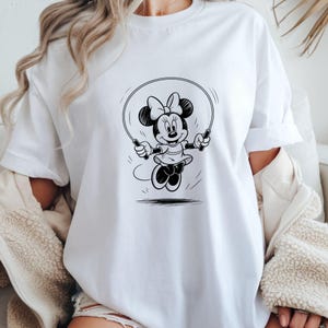 Puede incluir: Camiseta blanca con una ilustraci&oacute;n en blanco y negro de Minnie Mouse saltando a la cuerda. Minnie Mouse lleva un lazo, un vestido y zapatos. Un dise&ntilde;o de dibujos animados cl&aacute;sico.