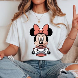 Puede incluir: Camiseta blanca con un dise&ntilde;o de dibujos animados de Minnie Mouse sosteniendo una taza de caf&eacute;. Minnie Mouse lleva un lazo y un vestido rosa. La imagen es un primer plano del dise&ntilde;o del personaje.