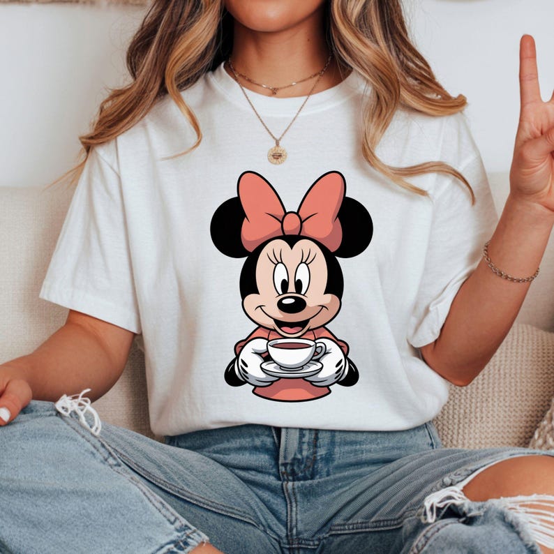 Puede incluir: Camiseta blanca con un dise&ntilde;o de dibujos animados de Minnie Mouse sosteniendo una taza de caf&eacute;. Minnie Mouse lleva un lazo y un vestido rosa. La imagen es un primer plano del dise&ntilde;o del personaje.