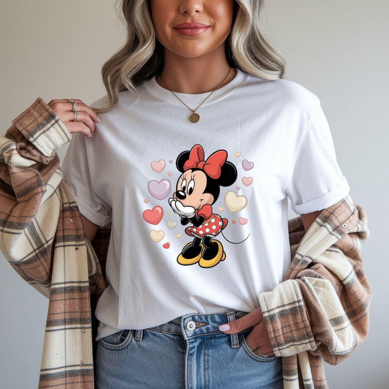 Op de afbeelding: Wit T-shirt met een Minnie Mouse cartoonontwerp. Minnie draagt een rode strik, een jurk met witte stippen en gele schoenen, omringd door pastelkleurige harten. Het shirt wordt gecombineerd met een blauwe jeans en een geruit overhemd.