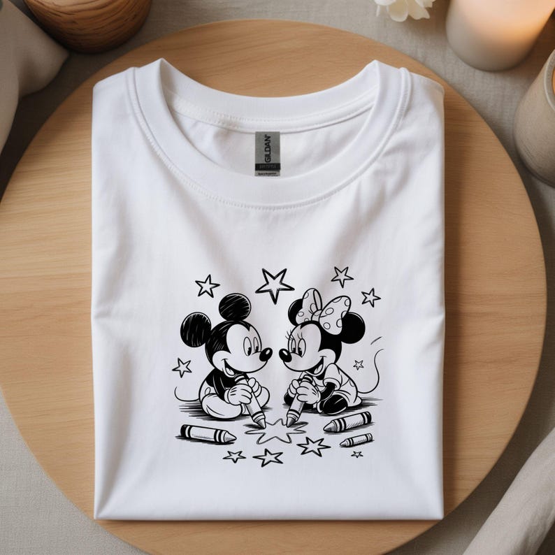 Puede incluir: Camiseta blanca con una ilustraci&oacute;n en blanco y negro de Mickey y Minnie Mouse dibujando estrellas con crayones. El dise&ntilde;o incluye estrellas y crayones. La camiseta est&aacute; doblada cuidadosamente sobre una superficie de madera.