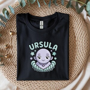 以下が含まれることがあります： 黒いTシャツ。漫画のイラストで、キャラクターの上に「URSULA」という名前があります。キャラクターは薄紫色で、星と貝殻のデザインに囲まれています。