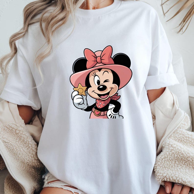May include: White t-shirt featuring a cartoon Minnie Mouse character in a pink cowgirl outfit, winking and holding a gold star. The design includes a pink hat, bow, and bandana. The shirt is a casual, everyday wear item.