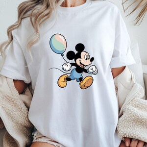 May include: White t-shirt featuring a cartoon illustration of Mickey Mouse holding a pastel-colored balloon. Mickey is depicted in his classic outfit: black shorts, yellow shoes, and white gloves. The design is centered on the shirt.