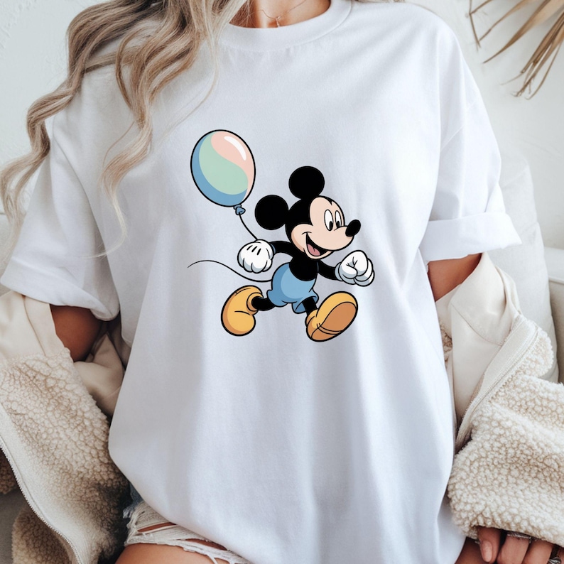 May include: White t-shirt featuring a cartoon illustration of Mickey Mouse holding a pastel-colored balloon. Mickey is depicted in his classic outfit: black shorts, yellow shoes, and white gloves. The design is centered on the shirt.