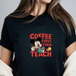May include: Black t-shirt with a cartoon Mickey Mouse design. Mickey is holding a coffee cup and a book. The red text reads "COFFEE FIRST, THEN TEACH". The design is centered on the shirt.