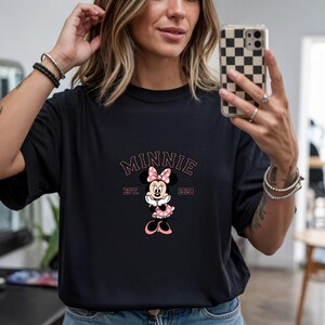 Może przedstawiać: Czarny t-shirt z grafiką Myszki Minnie. Projekt zawiera napis "MINNIE" w r&oacute;żowym łuku, tekst "EST. 1928" oraz postać Myszki Minnie w r&oacute;żowej sukience w kropki i kokardzie.