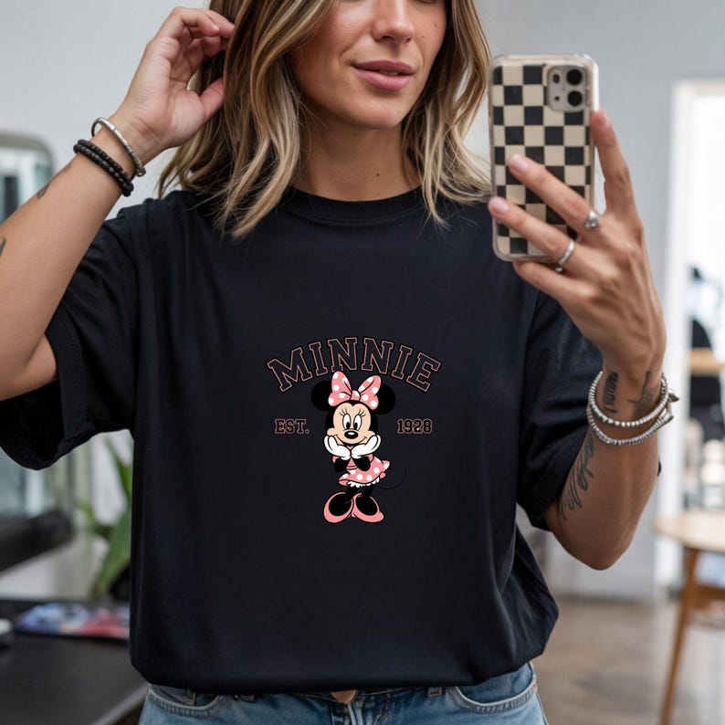 Może przedstawiać: Czarny t-shirt z grafiką Myszki Minnie. Projekt zawiera napis "MINNIE" w r&oacute;żowym łuku, tekst "EST. 1928" oraz postać Myszki Minnie w r&oacute;żowej sukience w kropki i kokardzie.