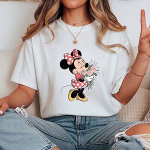 Op de afbeelding: Wit T-shirt met een Minnie Mouse-afbeelding. Minnie wordt afgebeeld in een rood-witte polkadotjurk en strik, met een boeket roze en paarse bloemen. Het shirt wordt gedragen met een blauwe spijkerbroek.