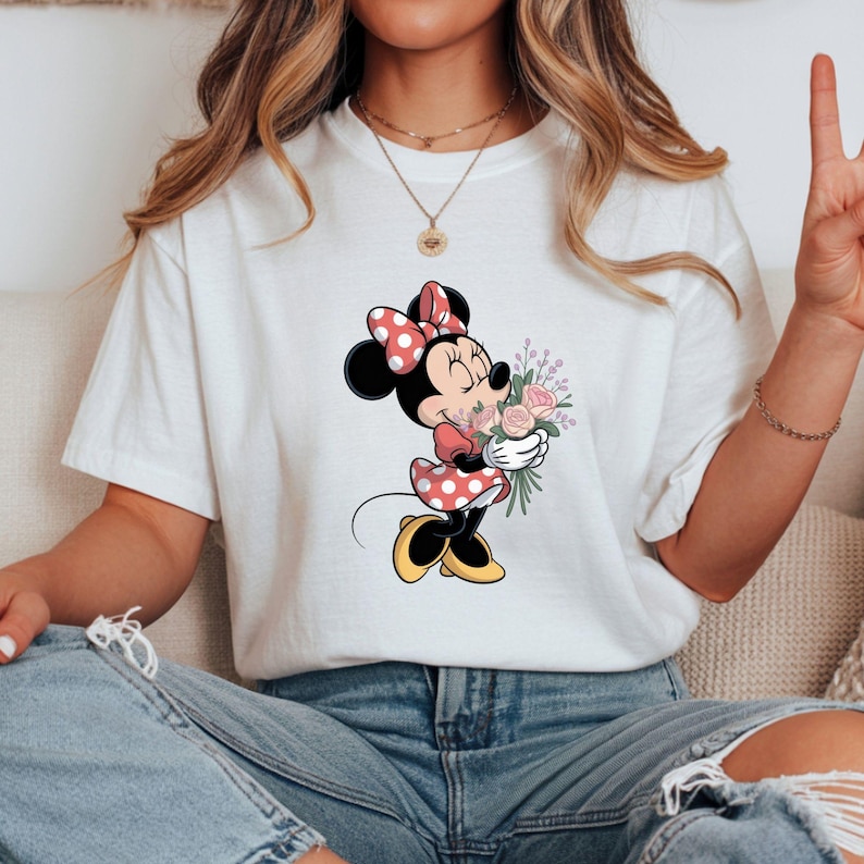 Op de afbeelding: Wit T-shirt met een Minnie Mouse-afbeelding. Minnie wordt afgebeeld in een rood-witte polkadotjurk en strik, met een boeket roze en paarse bloemen. Het shirt wordt gedragen met een blauwe spijkerbroek.