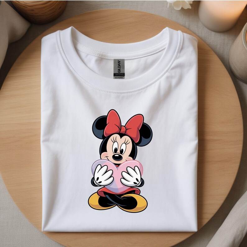 Op de afbeelding: Wit T-shirt met een Minnie Mouse-afbeelding. Het stripfiguur heeft een rode strik, zwarte oren en houdt een roze hart vast. Het T-shirt is op een houten oppervlak gevouwen.