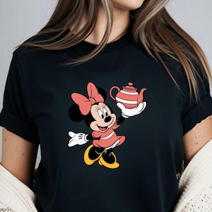 Puede incluir: Camiseta negra con un dise&ntilde;o de dibujos animados de Minnie Mouse sosteniendo una tetera a rayas rojas y blancas. Minnie lleva un vestido y un lazo rosas, zapatos amarillos y guantes blancos. Un dise&ntilde;o cl&aacute;sico de Disney.