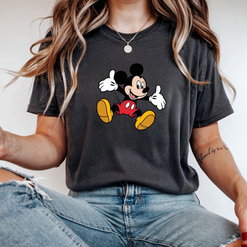 May include: Dark gray t-shirt featuring a cartoon image of Mickey Mouse. Mickey is depicted with black ears, a black face, white gloves, red shorts, and yellow shoes. The shirt is a casual style.