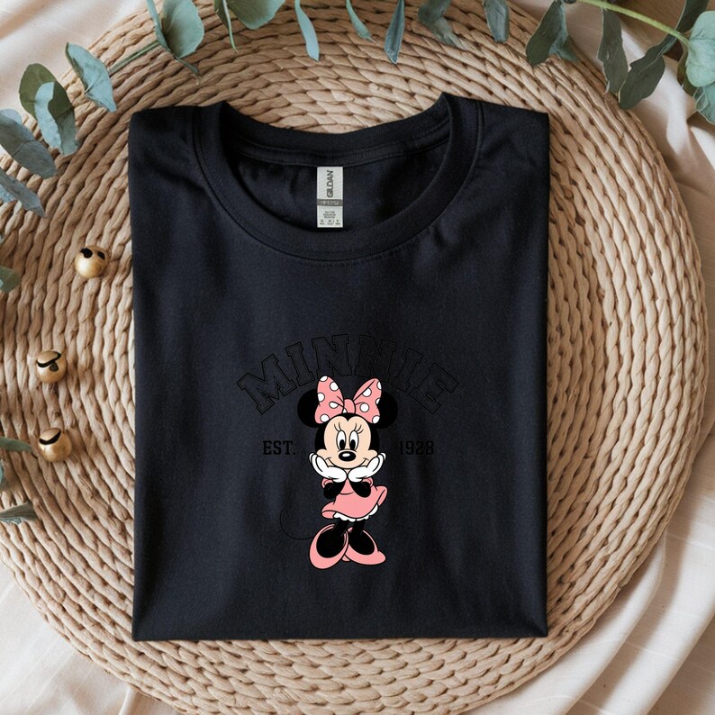 Może przedstawiać: Czarny t-shirt z grafiką Myszki Minnie. Projekt zawiera napis "MINNIE" nad wizerunkiem Myszki Minnie w r&oacute;żowej sukience i kokardce. Tekst "EST. 1928" znajduje się poniżej. Koszulka jest złożona na tkanej powierzchni.