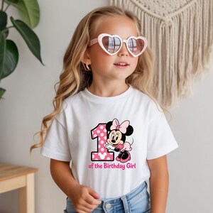 May include: A white t-shirt featuring a pink polka-dotted number 1 and a Minnie Mouse graphic. The text "of the Birthday Girl" is below the graphic. The girl is wearing heart-shaped sunglasses.