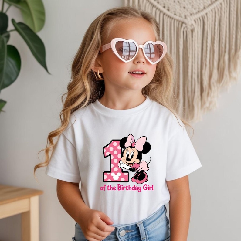 May include: A white t-shirt featuring a pink polka-dotted number 1 and a Minnie Mouse graphic. The text "of the Birthday Girl" is below the graphic. The girl is wearing heart-shaped sunglasses.
