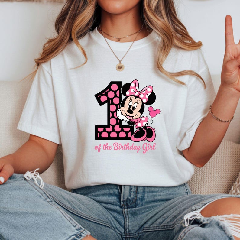 May include: White t-shirt featuring a graphic of Minnie Mouse with a pink polka dot bow and dress, and the number 1 with pink polka dots. Text below reads "of the Birthday Girl".