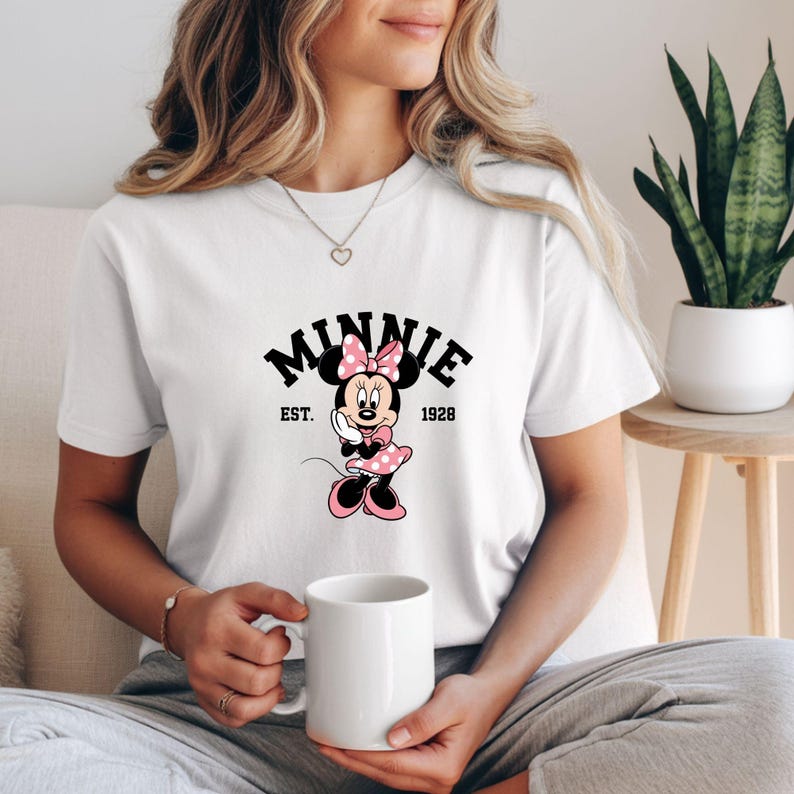 Może przedstawiać: Biała koszulka z grafiką Myszki Minnie. Projekt zawiera słowo "Minnie" w kolorze czarnym, z napisem "Est. 1928" poniżej. Myszka Minnie jest przedstawiona w kolorze r&oacute;żowym i czarnym, w sukience w kropki i kokardą.