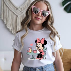 Puede incluir: Camiseta blanca con un dise&ntilde;o de dibujos animados de Minnie Mouse. Minnie est&aacute; sentada a una mesa con un p&aacute;jaro, sosteniendo una taza de t&eacute;. El dise&ntilde;o incluye detalles en rosa y negro, con un coraz&oacute;n encima de la mesa. La persona lleva gafas de sol rosas en forma de coraz&oacute;n.
