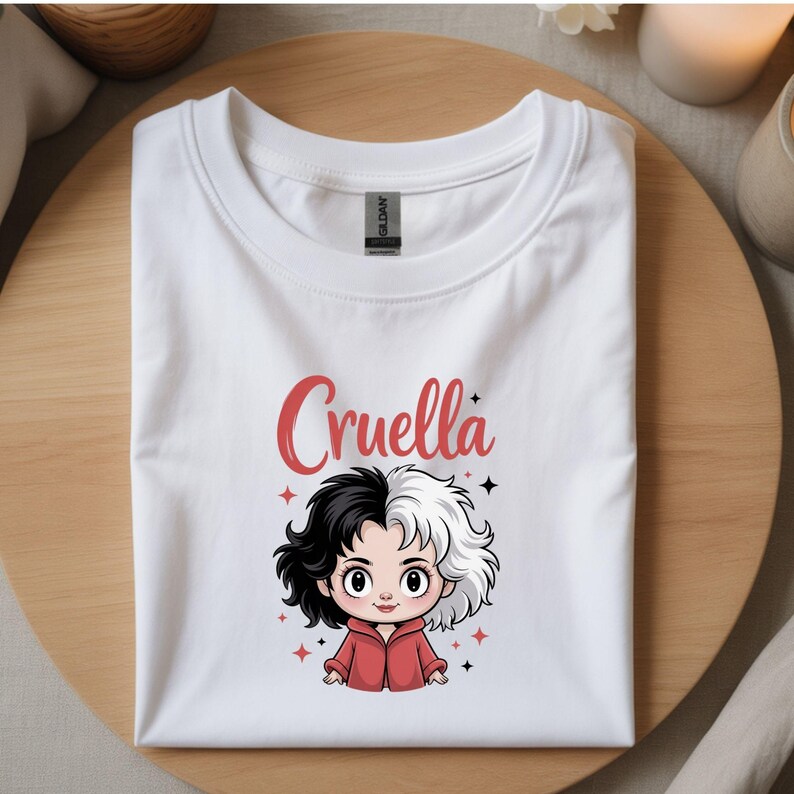 以下が含まれることがあります： 白のTシャツ。クルエラ・ド・ヴィルの漫画イラストが描かれており、黒と白の髪と赤いジャケットを着ています。「Cruella」という名前が赤い文字でキャラクターの上に書かれています。木製の表面に折りたたまれたTシャツ。