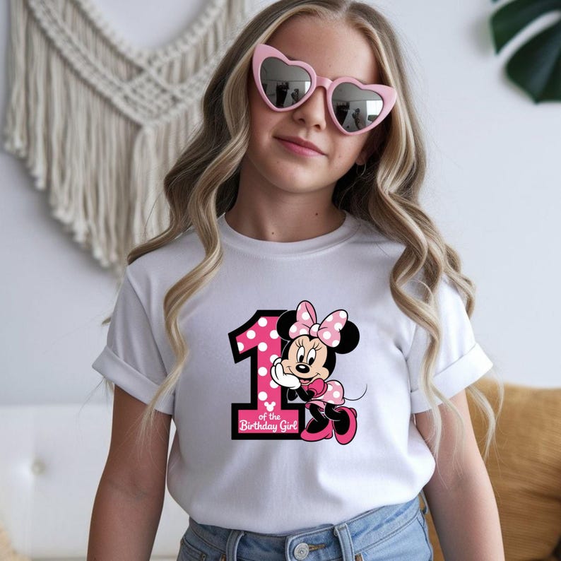May include: White t-shirt featuring a Minnie Mouse graphic with the number 1 in pink polka dots and the text "of the Birthday Girl". The shirt is worn by a person wearing pink heart-shaped sunglasses.