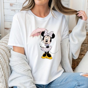 Op de afbeelding: Wit T-shirt met een Minnie Mouse-afbeelding. Minnie wordt afgebeeld met een roze paraplu, draagt een lavendelkleurige jurk met stippen, een bijpassende strik en gele schoenen. Het ontwerp is gecentreerd op het shirt.
