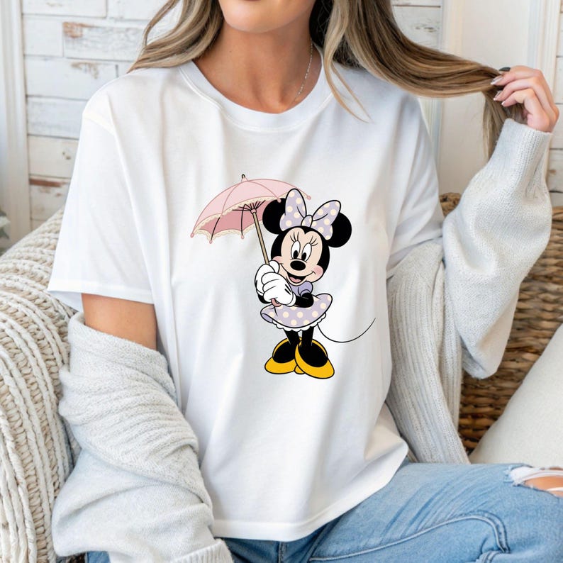 Op de afbeelding: Wit T-shirt met een Minnie Mouse-afbeelding. Minnie wordt afgebeeld met een roze paraplu, draagt een lavendelkleurige jurk met stippen, een bijpassende strik en gele schoenen. Het ontwerp is gecentreerd op het shirt.
