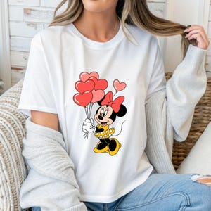 May include: White t-shirt featuring a cartoon Minnie Mouse holding heart-shaped balloons. Minnie Mouse is wearing a yellow dress with white polka dots and a red bow. The balloons are various shades of red and pink.