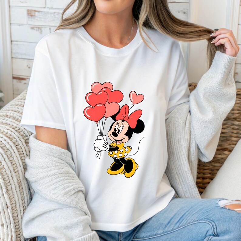 May include: White t-shirt featuring a cartoon Minnie Mouse holding heart-shaped balloons. Minnie Mouse is wearing a yellow dress with white polka dots and a red bow. The balloons are various shades of red and pink.