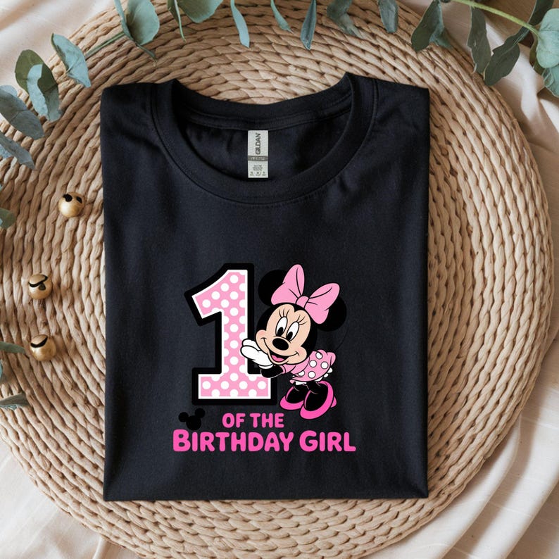 May include: Black t-shirt featuring a pink polka-dotted number 1 and a Minnie Mouse graphic with a pink bow and dress. The text "OF THE BIRTHDAY GIRL" is in pink. The shirt is folded neatly.